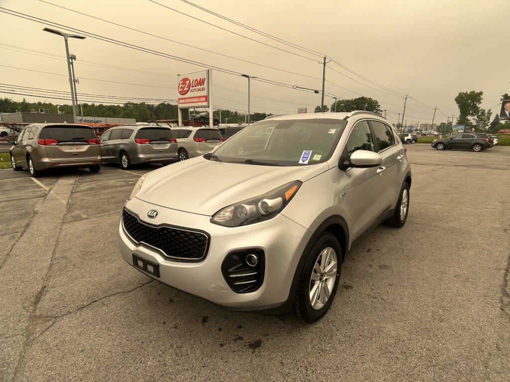 Silver Kia SUV in the foreground of a car dealership lot with several minivans parked nearby and an EZ Loan Auto Sales sign in the background, under overcast skies.