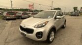 Silver Kia SUV in the foreground of a car dealership lot with several minivans parked nearby and an EZ Loan Auto Sales sign in the background, under overcast skies.
