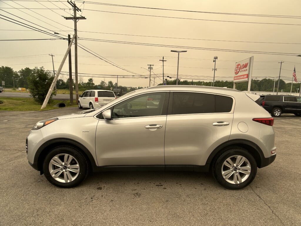 Silver Kia SUV in a dealership parking lot with overhead power lines and a dealership sign in the background.