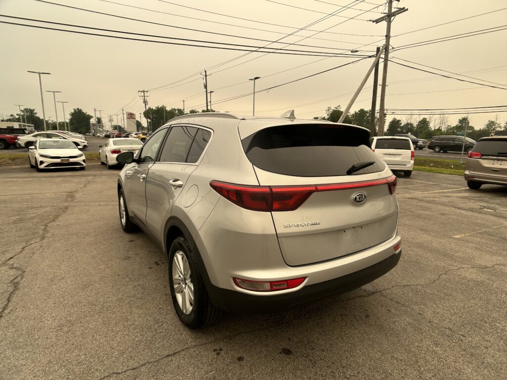 Rear three-quarter view of a silver Kia Sportage in a dealership lot with other cars and utility poles in the background to the right and overhead power lines.