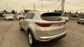 Rear three-quarter view of a silver Kia Sportage in a dealership lot with other cars and utility poles in the background to the right and overhead power lines.