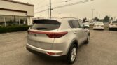 Rear three-quarter view of a white Kia SUV parked in a dealership lot with a building on the left and power lines overhead.