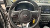 Driver's POV inside a Kia: steering wheel with Kia logo, dashboard gauges, and center console; outside a parking lot of cars through the windshield