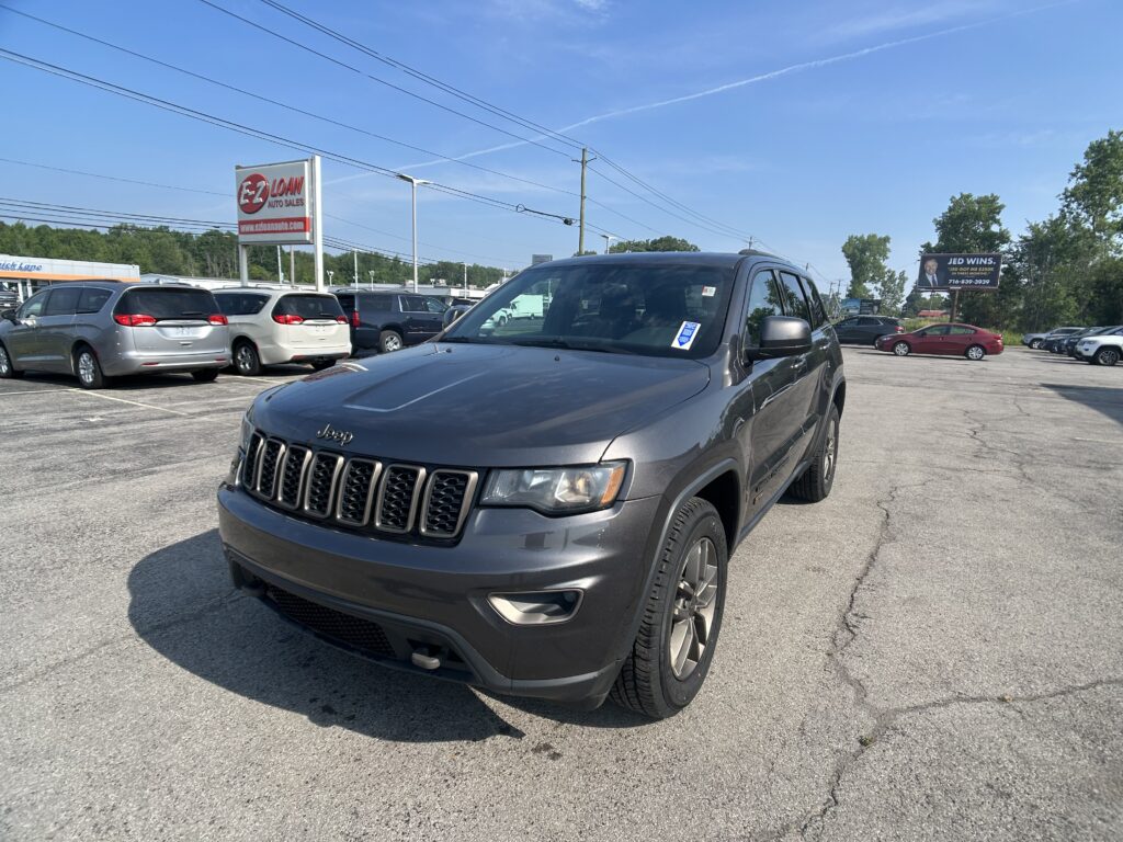Gray Jeep SUV in a car dealership lot with several minivans parked nearby and an EZ Loan Auto Sales sign in the background against a clear blue sky.