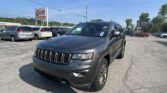 Gray Jeep SUV in a car dealership lot with several minivans parked nearby and an EZ Loan Auto Sales sign in the background against a clear blue sky.