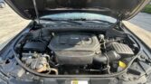 Open car hood revealing the engine bay with a V6 engine cover, battery on the left, and various hoses and components visible beneath the hood panel.