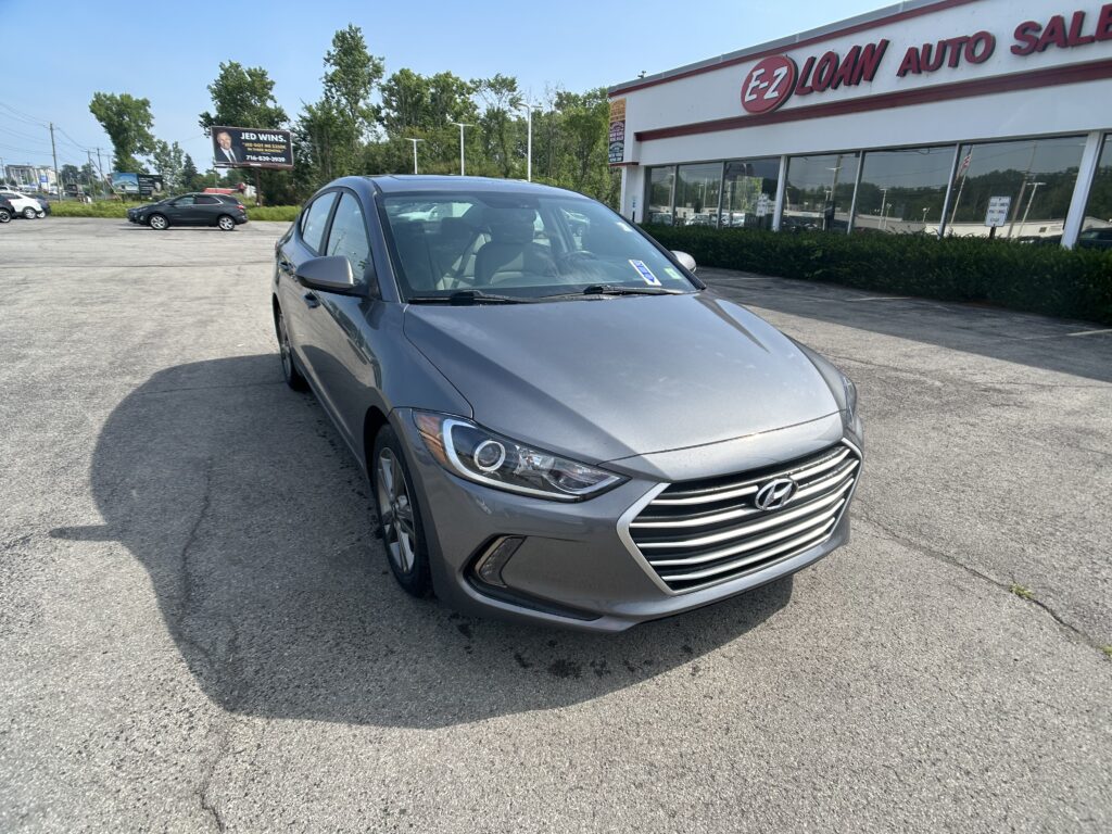 Silver Hyundai sedan parked in a lot in front of an EZ Loan Auto Sales storefront with large glass windows and bushes nearby.