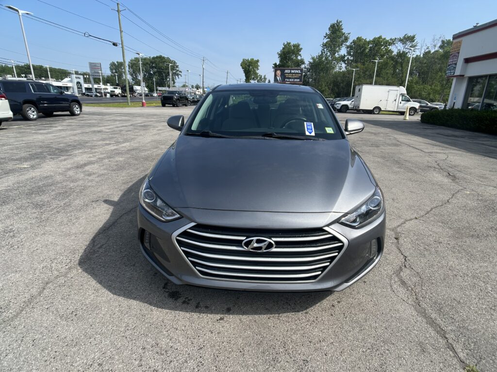 Front view of a gray Hyundai sedan parked in a crowded lot with dealership buildings in the background.