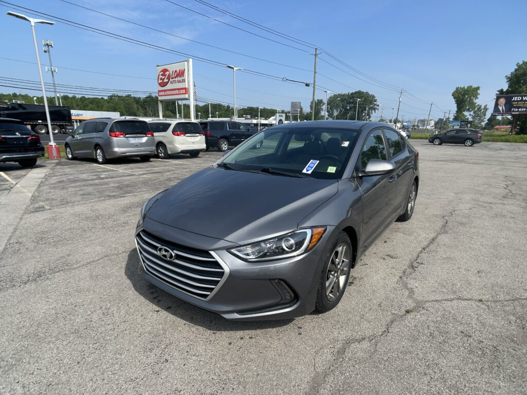 Gray Hyundai sedan in a crowded parking lot with other vehicles, billboards, and power lines under a clear blue sky.
