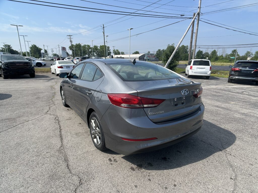 Gray Hyundai Elantra seen from the rear in a parking lot with other cars and a blue sky above, power lines overhead.