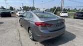 Gray Hyundai Elantra seen from the rear in a parking lot with other cars and a blue sky above, power lines overhead.