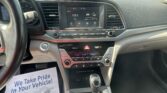 Car interior showing the dashboard and center touchscreen displaying 106.5 FM, date and time, with gear selector and climate controls visible; papers on the passenger side say 'We Take Pride In Your Vehicle.'