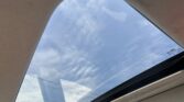 View through a car's large panoramic sunroof looking up at a partly cloudy blue sky.