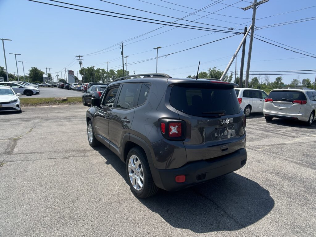 Rear three-quarter view of a dark gray Jeep SUV parked in a crowded car lot under overhead power lines on a sunny day.