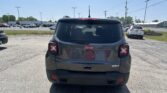 Rear view of a dark gray Jeep Latitude SUV in a sunny parking lot with other cars and streetlights in the background, tailgate visible with Latitude badge.