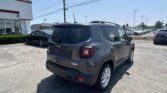 Rear view of a gray Jeep Latitude SUV parked in a dealership lot with rows of cars and a white building nearby in the background.