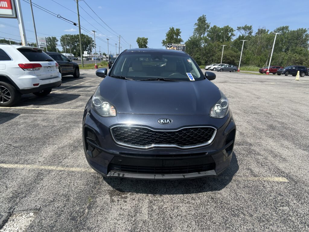 Front view of a dark blue Kia SUV parked in a busy car lot with other cars and a row of trees in the background.