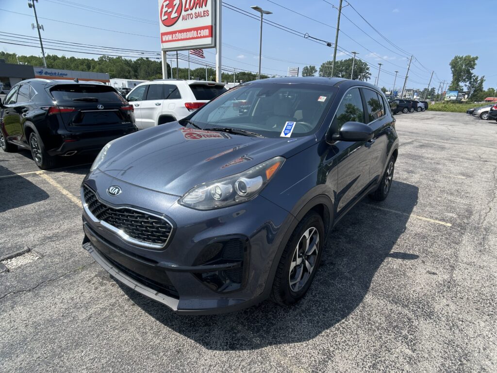 Close-up of a blue Kia SUV in a car dealership lot with other vehicles and a tall EZ Loan Auto Sales sign in the background, under a clear sky.