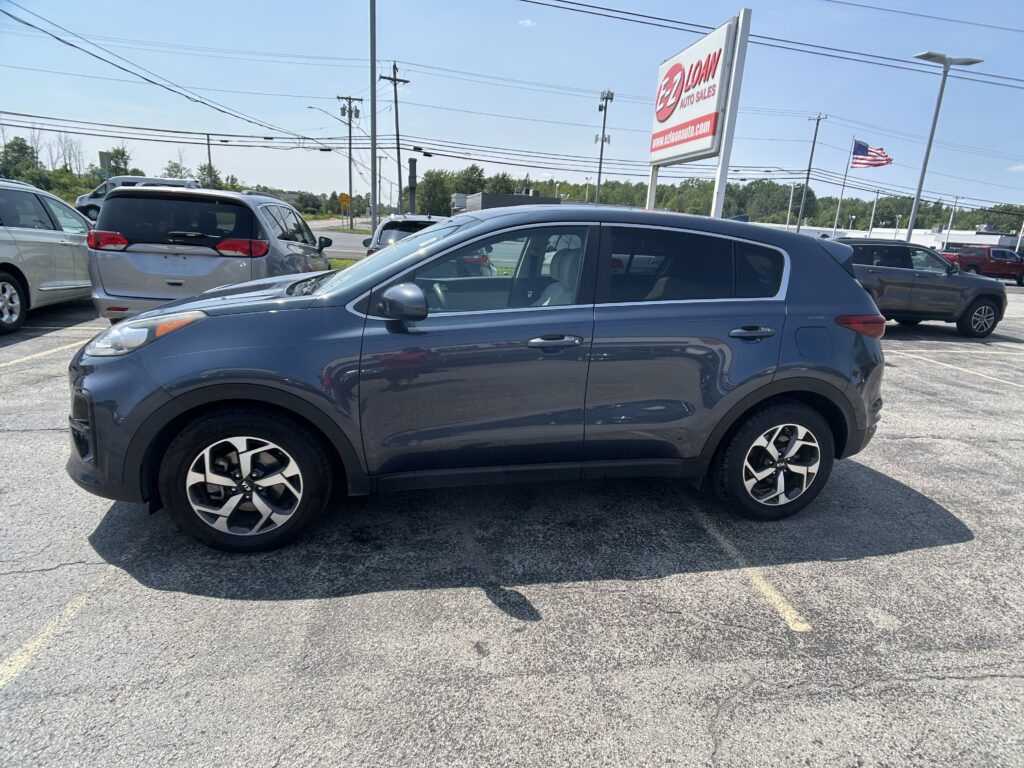 Blue SUV in a car dealership lot with other vehicles and a tall Auto Sales sign; American flag in the background.