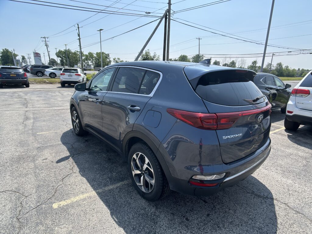Rear three-quarter view of a dark gray Kia Sportage parked in a lot with other cars and power lines overhead on a sunny day.