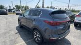 Rear three-quarter view of a dark gray Kia Sportage parked in a lot with other cars and power lines overhead on a sunny day.