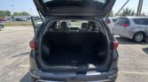 Open rear hatch of a dark SUV in a parking lot, showing a large empty cargo area and visible back seats. Behind it are other parked cars and a bright daytime sky.