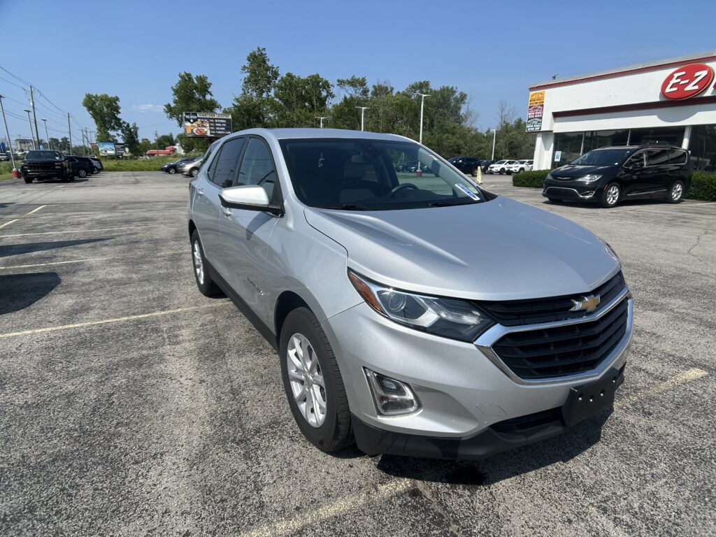 Front-right view of a silver Chevrolet SUV parked in a lot, with a white building and an E-Z sign on the right and other cars in the background.