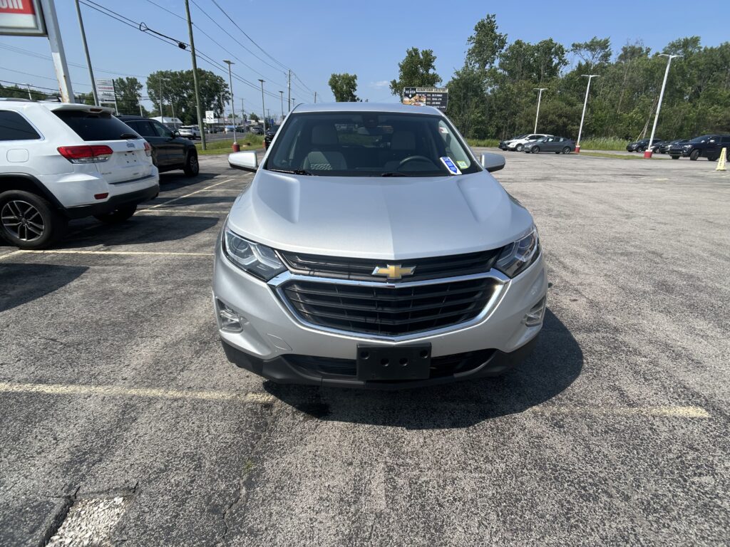 Front view of a silver Chevrolet SUV in a parking lot with other vehicles and a clear blue sky in the background.