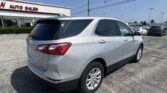 Rear three-quarter view of a silver SUV parked in a dealership lot near a white building with 'AUTO SALES' in red letters against a blue sky and power lines above.