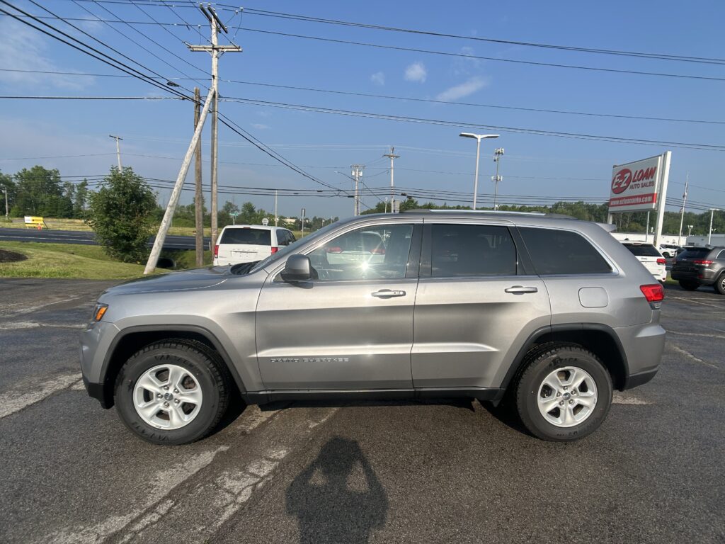 Silver SUV (Jeep Grand Cherokee) in a dealership lot with power lines overhead and an EZ Loan Auto Sales sign in the background.