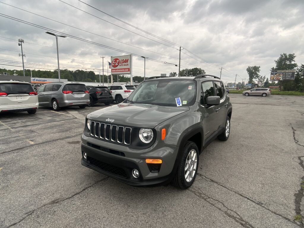 Gray Jeep SUV in the foreground of a car dealership lot, with a red-and-white EZ LOAN Auto Sales sign and several other vans in the background, under a cloudy sky.