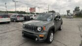 Gray Jeep SUV in the foreground of a car dealership lot, with a red-and-white EZ LOAN Auto Sales sign and several other vans in the background, under a cloudy sky.
