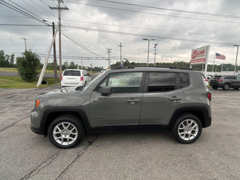 Gray Jeep Renegade SUV parked in foreground at a car dealership lot under cloudy skies, with an EZ Loan Auto Sales sign and American flag in the background.