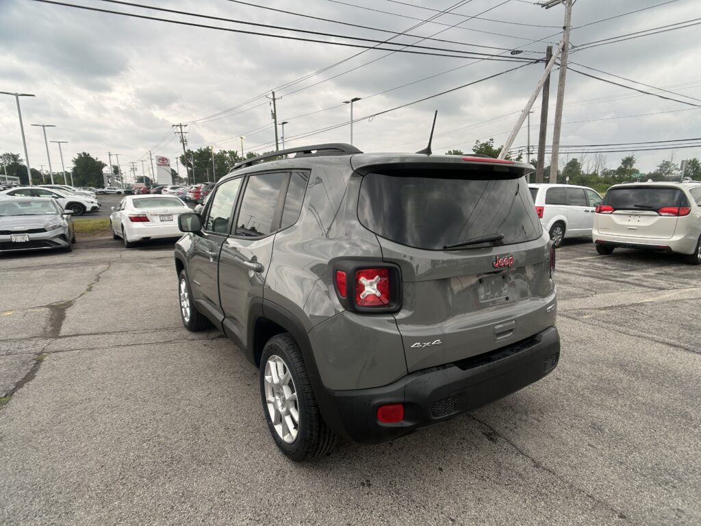 Rear three-quarter view of a gray Jeep SUV in a car dealership lot with other vehicles and a Toyota sign in the distance.