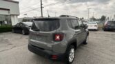 Rear view of a gray Jeep SUV in a dealership parking lot with other cars and a white building on the left under a cloudy sky.
