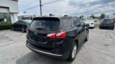 Rear three-quarter view of a black Chevrolet SUV in a car dealership lot, with other cars and a Toyota dealership sign in the background under a cloudy sky.