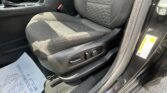Car interior focusing on the front seat with textured fabric, showing the seat adjustment lever, center console, and a red seat belt buckle; a paper lies on the floor mat nearby.