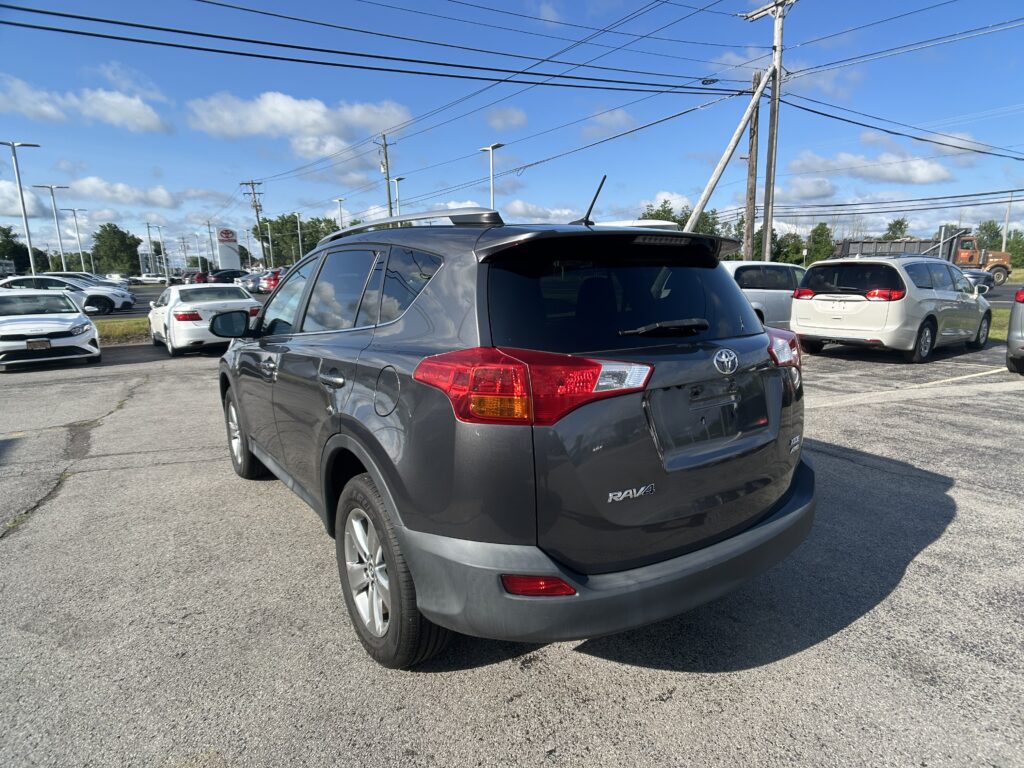 Rear three-quarter view of a gray Toyota RAV4 in a car dealership lot, other vehicles visible around, under a blue sky with power lines overhead.