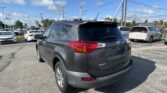 Rear three-quarter view of a gray Toyota RAV4 in a car dealership lot, other vehicles visible around, under a blue sky with power lines overhead.