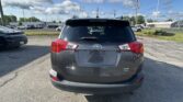 Rear view of a gray Toyota RAV4 XLE AWD parked in a car lot, with a black car to the left and dealership buildings in the background.