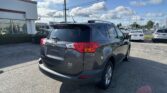 Rear three-quarter view of a gray Toyota RAV4 in a dealership parking lot, with a white building on the left and power lines overhead under a blue sky with clouds.