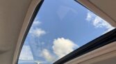 Interior view from a car showing a large open sunroof with a clear blue sky and white clouds outside, beige headliner surrounding the glass panel