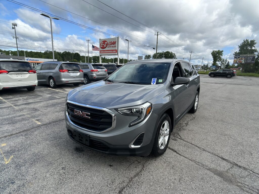 Silver GMC SUV in the foreground at an EZ Loan Auto Sales lot, with other vehicles and signs in the background under a cloudy sky.