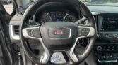 Driver's perspective of a GMC car interior: steering wheel, instrument cluster, and center touchscreen visible; gear selector in Park, parked cars outside through the windshield.