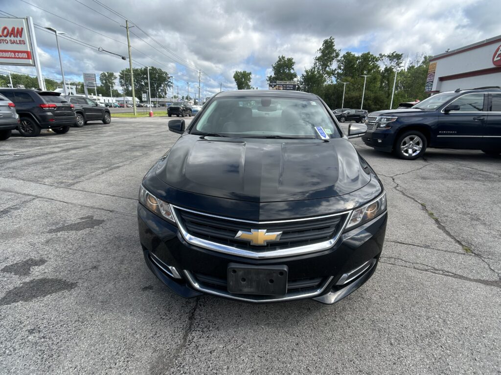 Front view of a dark Chevrolet car in a dealership lot with other vehicles and a building in the background