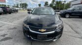 Front view of a dark Chevrolet car in a dealership lot with other vehicles and a building in the background