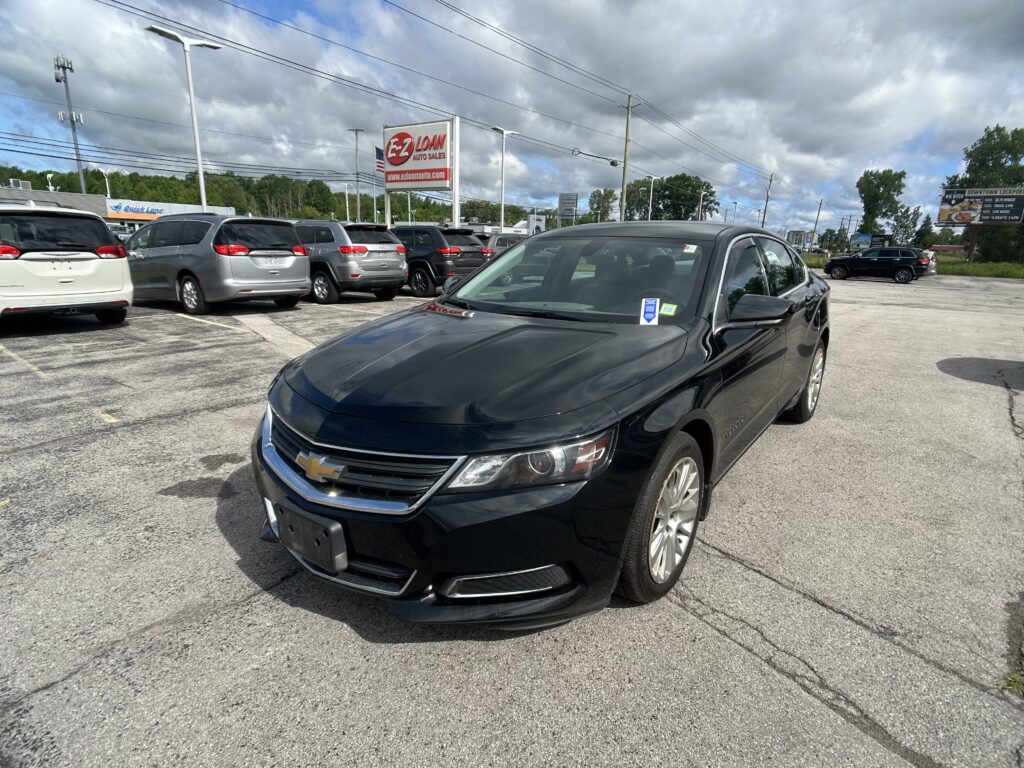Black Chevrolet sedan in the foreground of a dealership lot with silver minivans and an EZ Loan Auto sign in the background under a cloudy sky.