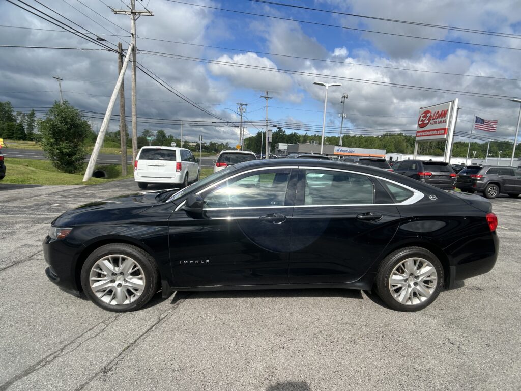 Black Chevrolet Impala in a car dealership lot, with an EZ LOAN Auto Sales sign and an American flag in the background under a cloudy sky.