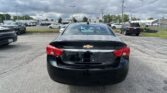 Rear view of a black Chevrolet sedan with a V6 badge, parked in a busy car lot on a cloudy day.