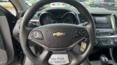 Driver's view of a Chevrolet steering wheel with dashboard gauges and center touchscreen in a car showroom lot backdrop outside the windshield